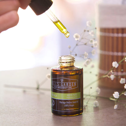 HILLS GARDEN 100% pure cold-pressed moringa oil in a 30ml amber glass dropper bottle with a hand applying a drop.